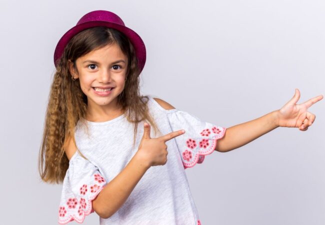 smiling-little-caucasian-girl-with-purple-party-hat-pointing-side-isolated-white-wall-with-copy-space